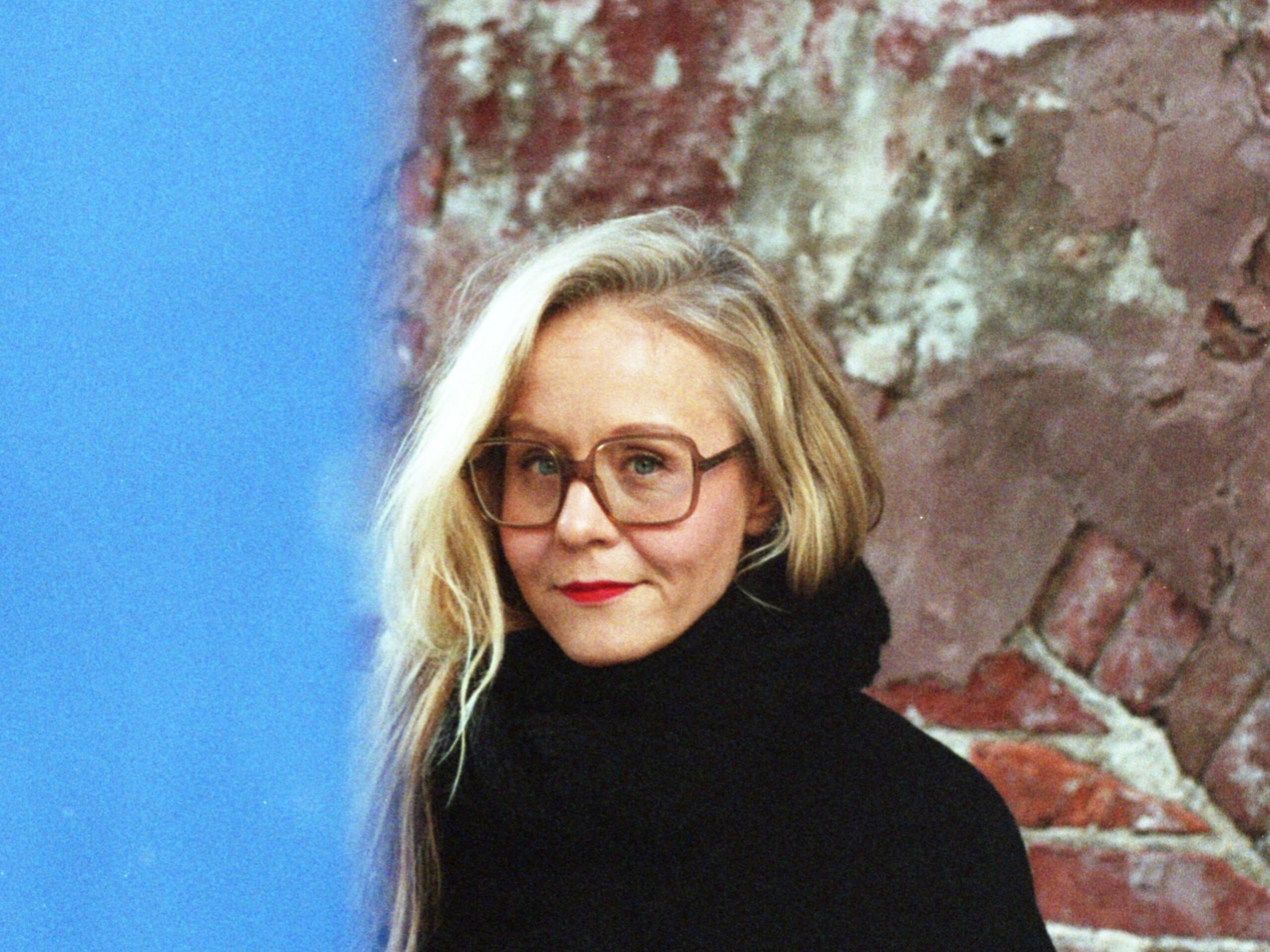 Anna-Kaari Hakkarainen katsoo kameraan. Seisoo punaista tiiliseinää vasten.